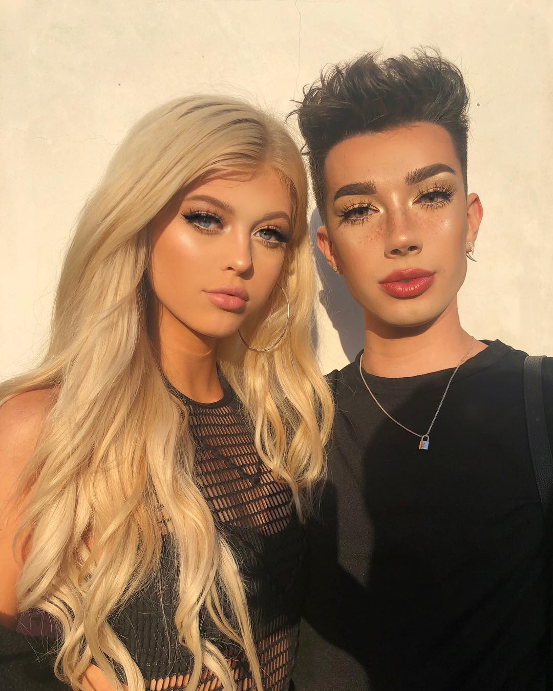 a photograph of two people standing sidebyside, posing for the camera. the person on the left has long blonde hair, hoop earrings, and is wearing a black mesh top. the person on the right has dark hair, freckles, is wearing makeup, and has a necklace with a small padlock pendant. both individuals are wearing dark shirts.