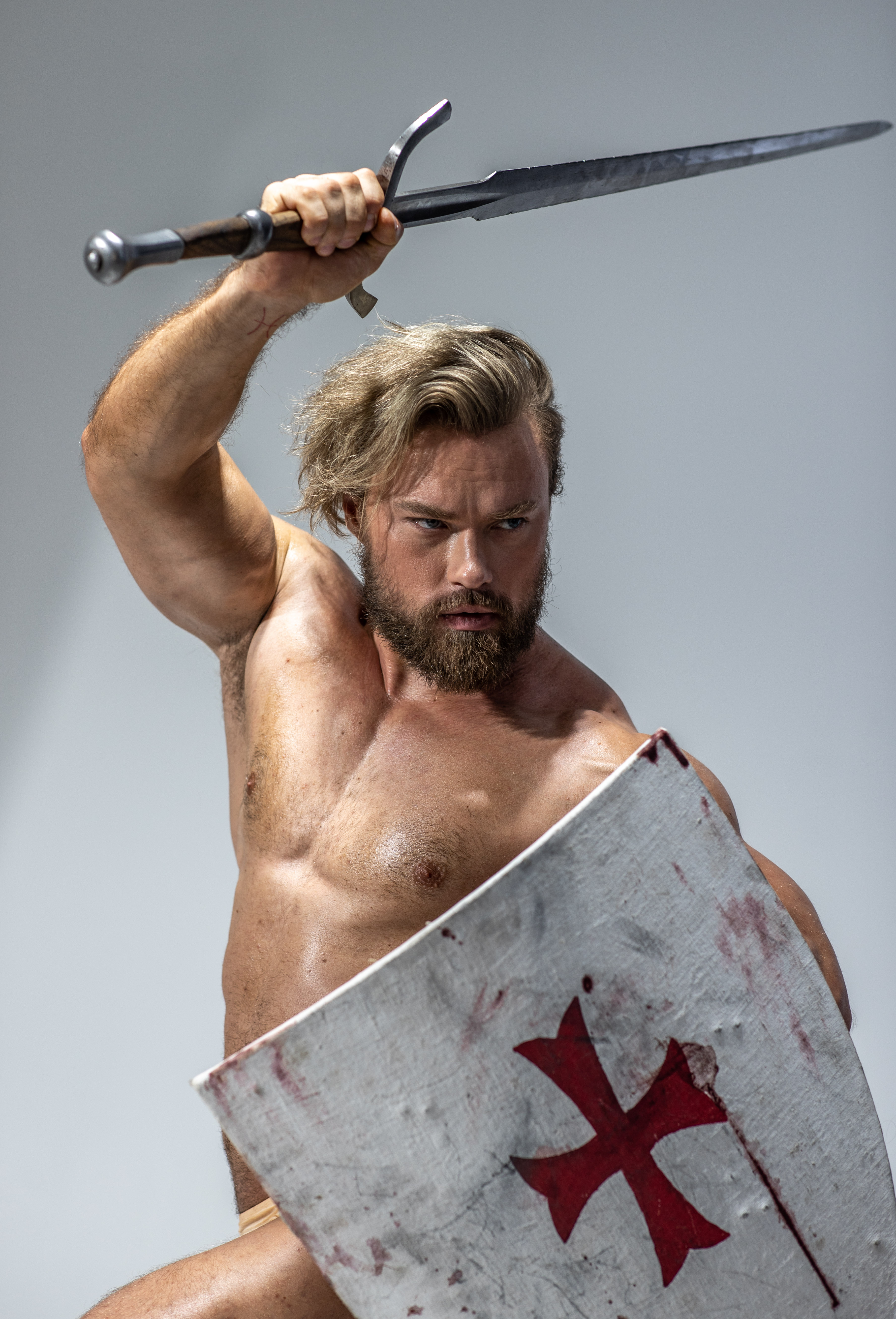 a muscular man, stripped to the waist, embodies a fierce warrior. he holds a gleaming sword aloft in his right hand, ready to strike. his left hand grips a large, battered shield emblazoned with a bloodred cross. his face, framed by a blonde beard, is a mask of fierce determination. the studio lighting emphasizes his powerful physique and the dramatic nature of the scene.
