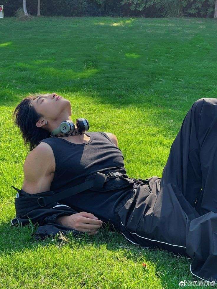 a man in a black shirt and pants is laying on a grassy field in a park, looking up at the sun. he has headphones around his neck and is wearing a black jacket over his shoulders.  there are trees in the background, out of focus.