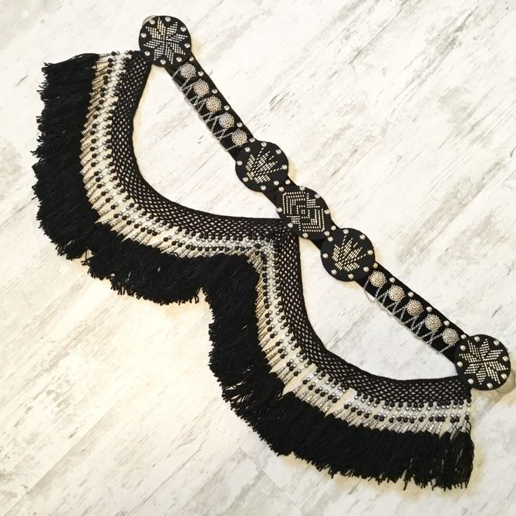 a black and silver beaded belt with fringe is displayed on a wooden surface. the belt is elaborately decorated with intricate patterns and beadwork. the fringe adds a dramatic touch. the overall aesthetic is ornate and eyecatching.