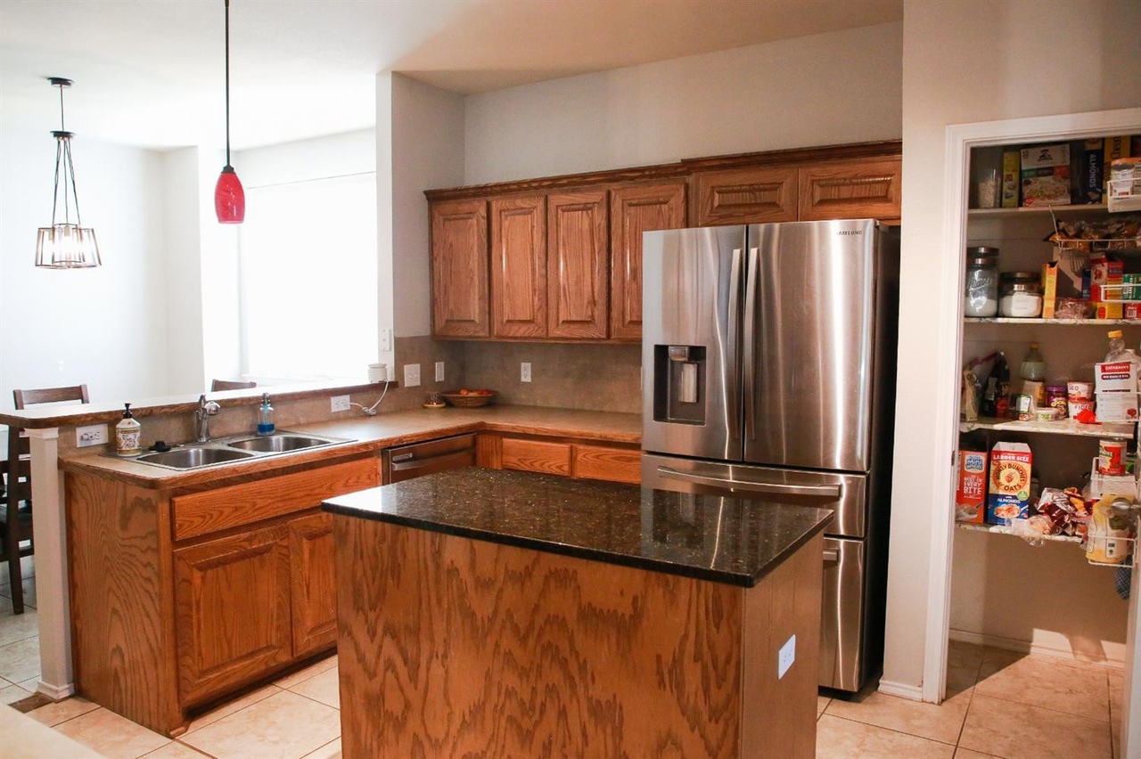 this image shows a kitchen with brown cabinets and a countertop, stainless steel refrigerator, kitchen island, stainless steel sink, and a pantry with a variety of food items. there are also two light fixtures in the kitchen.