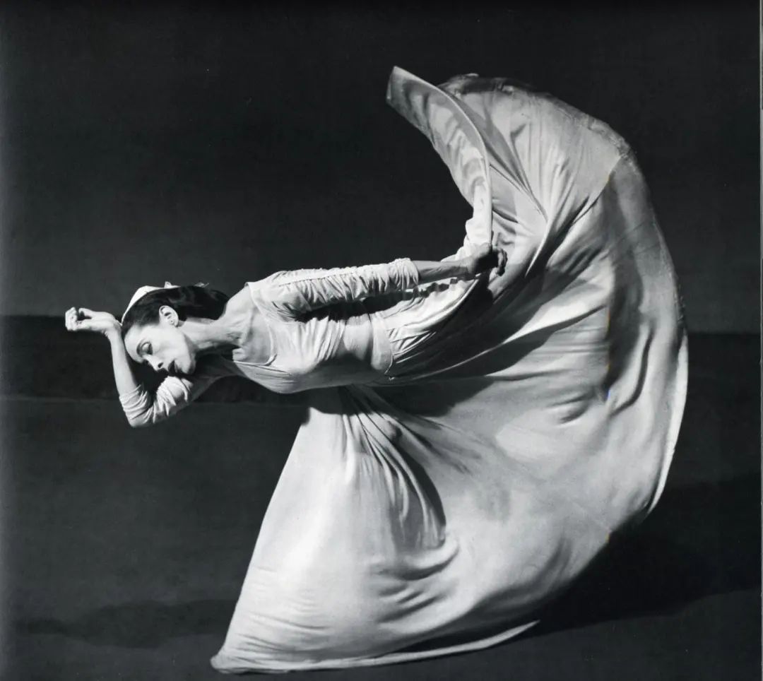 a black and white photograph of a dancer in a white dress, posing gracefully on a stage. the dancer's body is contorted in a dynamic pose, with her arms outstretched and her head tilted back. the fabric of her dress flows around her, creating a sense of motion and energy. the background is dark and blurry, drawing attention to the dancer's form. the photograph captures the beauty and artistry of dance, conveying a sense of power, grace, and emotion.  the photograph is likely a professional portrait of a dancer performing on stage.