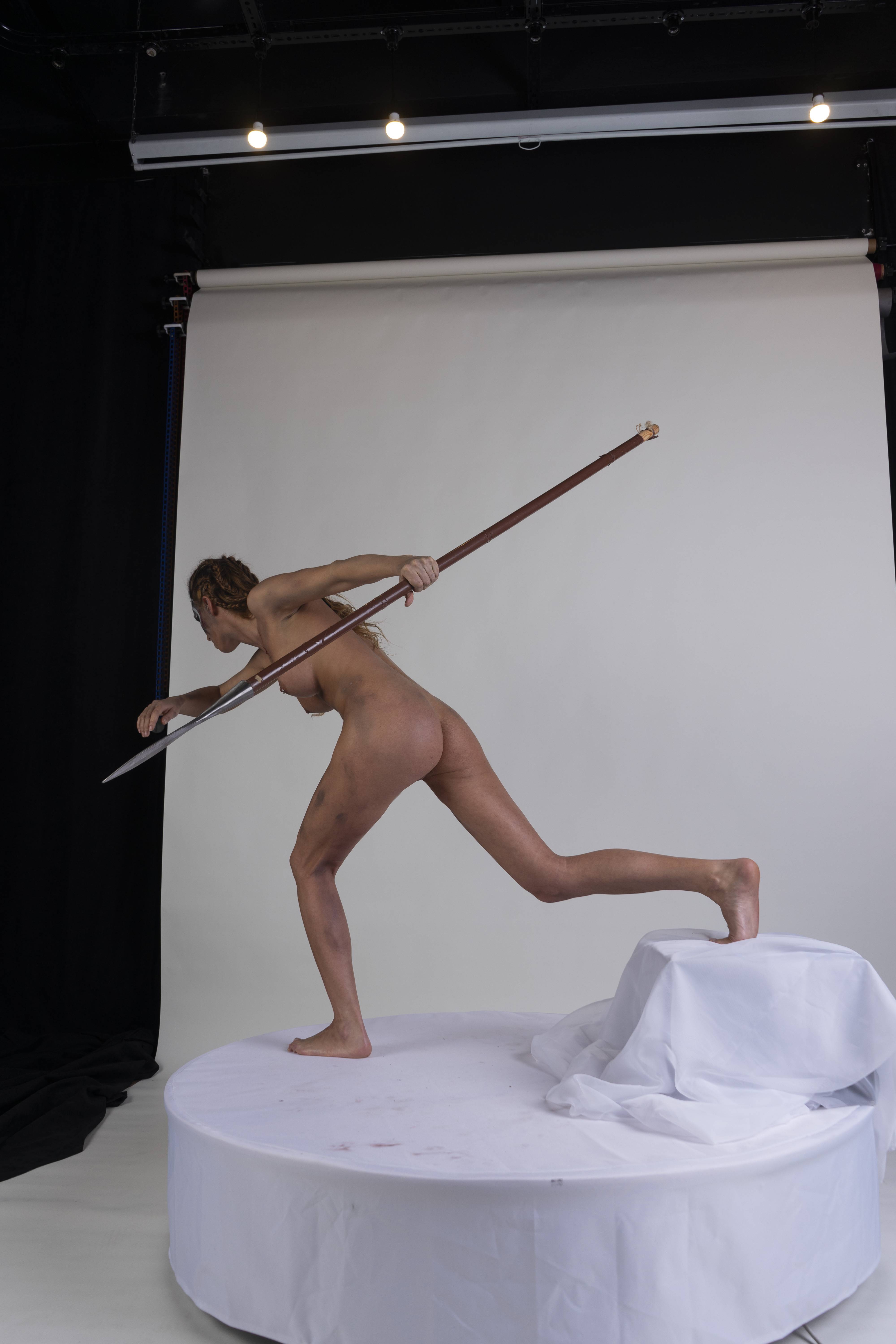 a full shot of a nude woman in a fighting stance, holding a spear. she is shown in profile view in a studio setting with a white background. she is standing on a white platform with draped fabric and is barefoot.