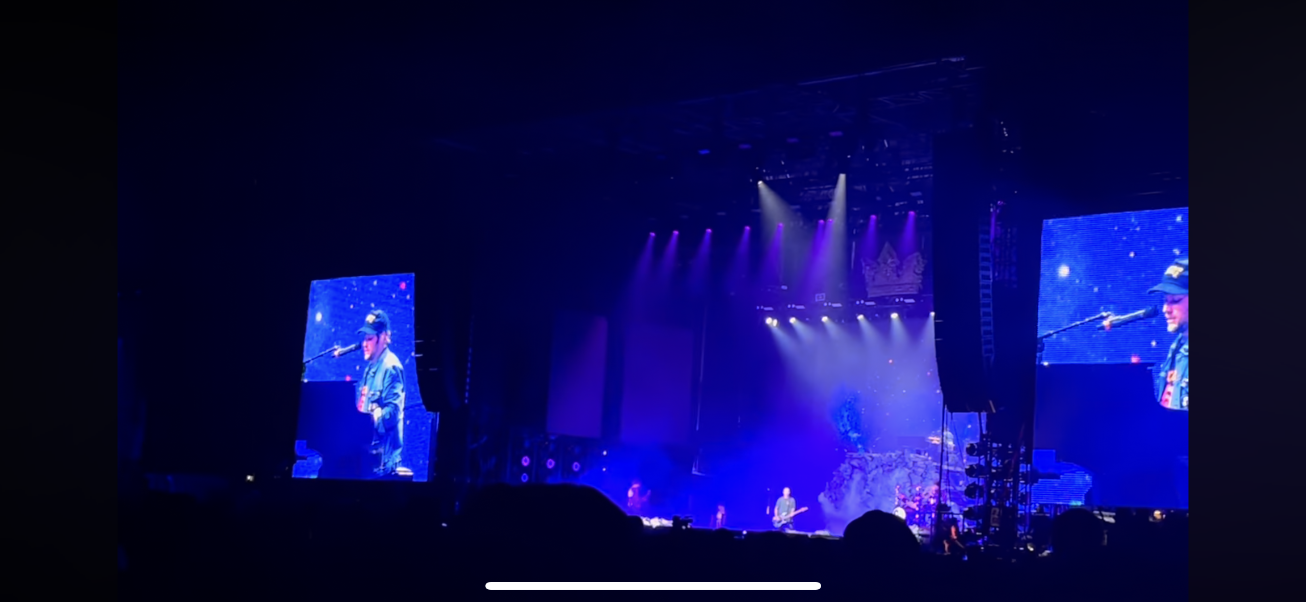 a concert stage is lit up with purple and blue spotlights, revealing a large video screen displaying a singer on stage. the singer is performing in front of a microphone with a black guitar strapped to their back. there is a crowd of people in the audience but they are mostly obscured by the darkness.  this image is a view of the stage at a concert.
