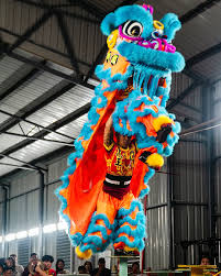 a colorful chinese lion dance performer is captured midair, showcasing the vibrancy and energy of this traditional performance. the performer, dressed in elaborate orange and blue costume, skillfully manipulates the lion's head and body, creating a sense of life and movement. the image, taken from a low angle, emphasizes the height and dynamism of the leap. the setting appears to be a large indoor space, possibly a warehouse or gymnasium, with a corrugated metal roof and a gathering of spectators in the background.