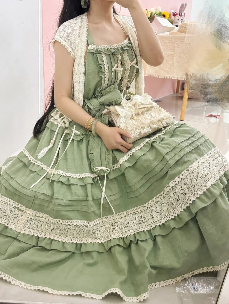 a young woman poses indoors, showcasing a stunning green lolita fashion dress. the dress features intricate lace detailing, delicate ruffles, and a tiered skirt. her romantic style evokes a charming, cottagecore aesthetic.