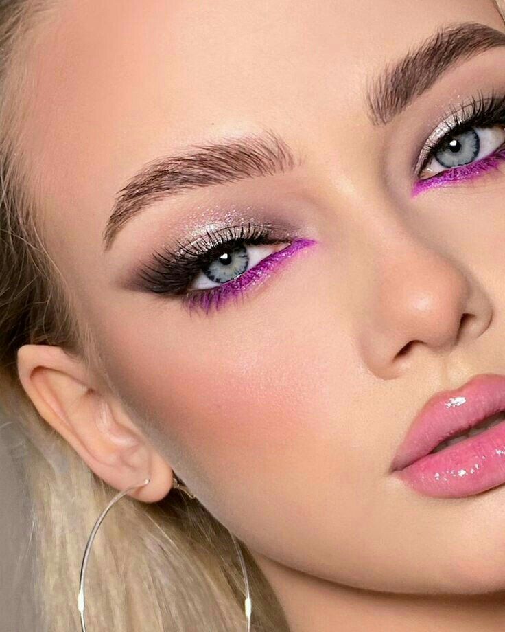 this is a closeup portrait of a woman showcasing her striking eye makeup. she has light, fair skin, blonde hair, and wears a large silver hoop earring. her eyebrows are welldefined and thick. her eye makeup includes silver eyeshadow, thick black eyeliner, long lashes, and a vivid pink eyeliner along her lower lash line. her lips are glossy and a bright pink color.