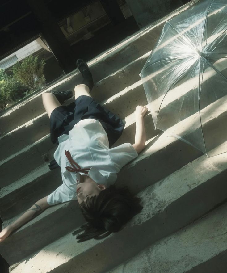 a young woman in a japanese school uniform with a long dark ponytail lies on a concrete staircase. she is wearing a white blouse with a red ribbon and a black skirt. an umbrella lies next to her. she is wearing black shoes. there is a shadow on the stairs behind her and she looks to be in a dark building outdoors.