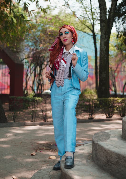 a stylish woman with vibrant red hair and striking makeup is captured in a fullbody outdoor shot. she is wearing a light blue pantsuit, a patterned necktie, and suspenders. her outfit is complemented by dangling earrings. the backdrop features a park setting with lush green bushes and trees, contrasting against a brightly painted wall in the background.  the woman is standing on a stone stairway, confidently posing for the camera. the overall vibe is fashionable and selfassured.