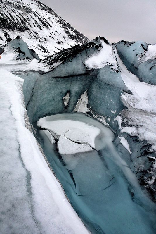 the image shows a glacier with deep crevasses, covered in snow and ice. the glacial ice has a blue hue. in the background, a snowcovered mountain can be seen. the sky is overcast.