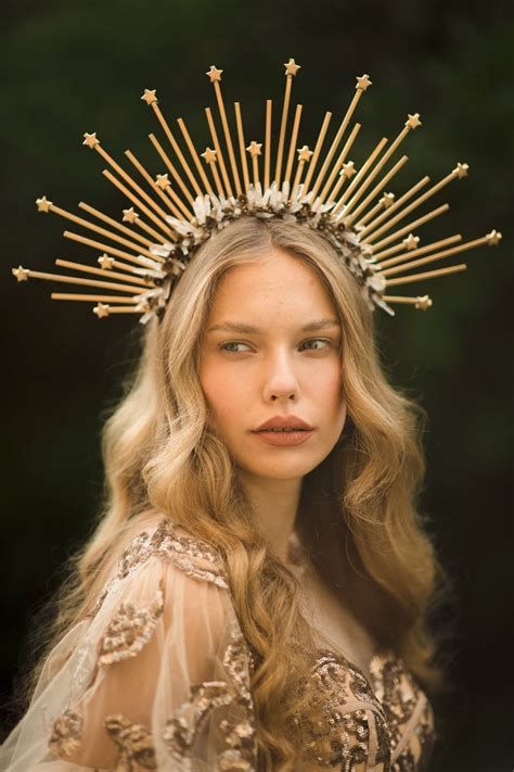 a woman with blonde hair and a golden crown adorned with stars poses in a natural setting. she wears a sheer, embellished dress. the lighting is soft and natural, highlighting her beauty and the ethereal quality of the image.