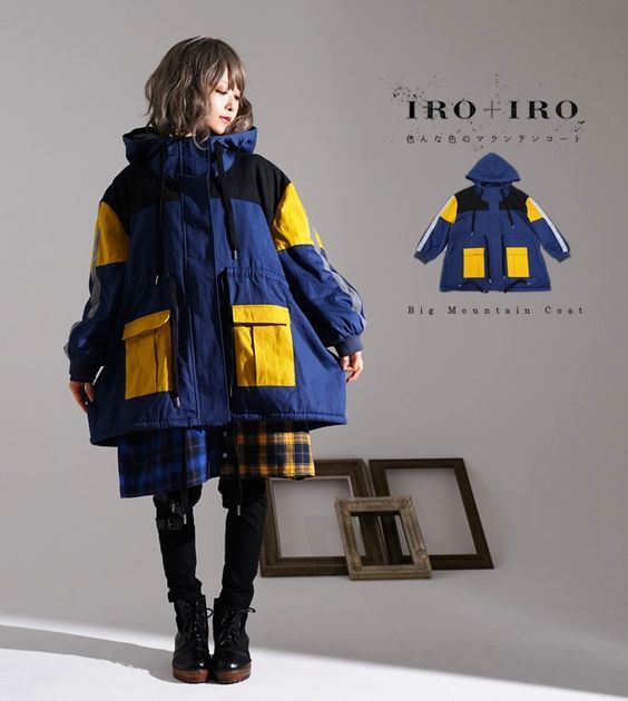 a woman is standing in a studio wearing a blue and yellow oversized coat with black detailing. it appears to be a puffer coat style, and she is wearing black leggings underneath, along with a plaid skirt.  she is wearing black boots and has short brown hair. the coat appears to be a big mountain coat and it has a brand name on it, "iro+iro." the background has a few wooden picture frames in it.