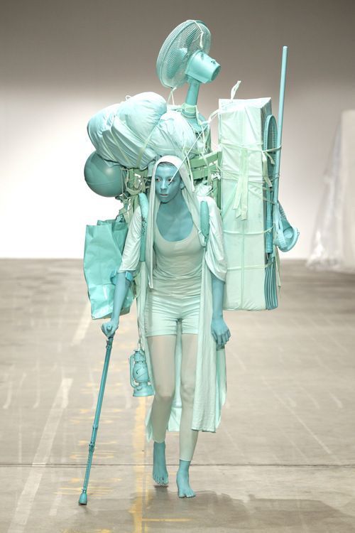 a model walks the runway during a fashion show.  they are wearing a blue and white outfit and carrying several objects that appear to be stacked on their back. their face is painted blue, and they are barefoot.  the runway and the background are a light gray.