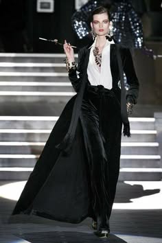 a woman wearing a chic black suit with a flowing coat stands on a runway, holding a cigarette in her hand, with a sophisticated and confident expression. her look is stylish and elegant, with a touch of vintage flair. she embodies a classic sense of fashion, making a statement with her tailored outfit and commanding presence.