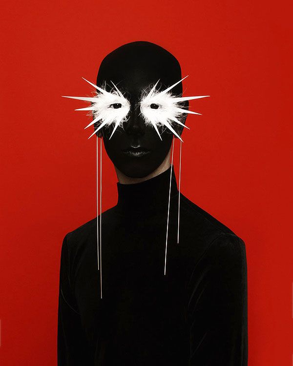 a person in a black turtleneck stands against a red background. their face is painted black and they are wearing a white, geometric mask with long, white, dangling strings that hang down to their chest.