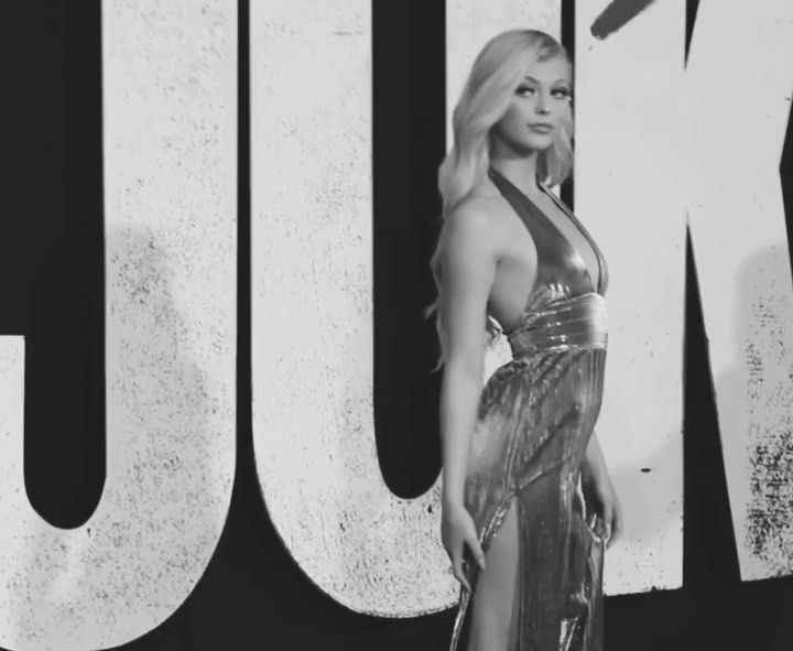 a woman with blonde hair stands in front of a backdrop wearing a halterneck evening gown with a slit in the skirt, captured in black and white.