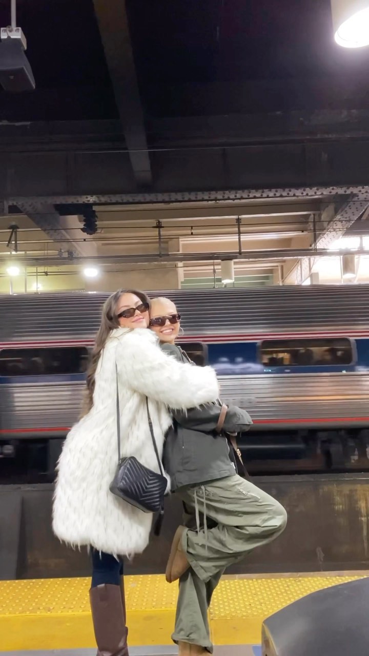 two women are hugging at a train station. the woman on the left is wearing sunglasses, a white faux fur coat, blue jeans, and brown boots. she has long brown hair. the woman on the right is wearing sunglasses, a gray jacket, and loose green pants. there is a train behind them.