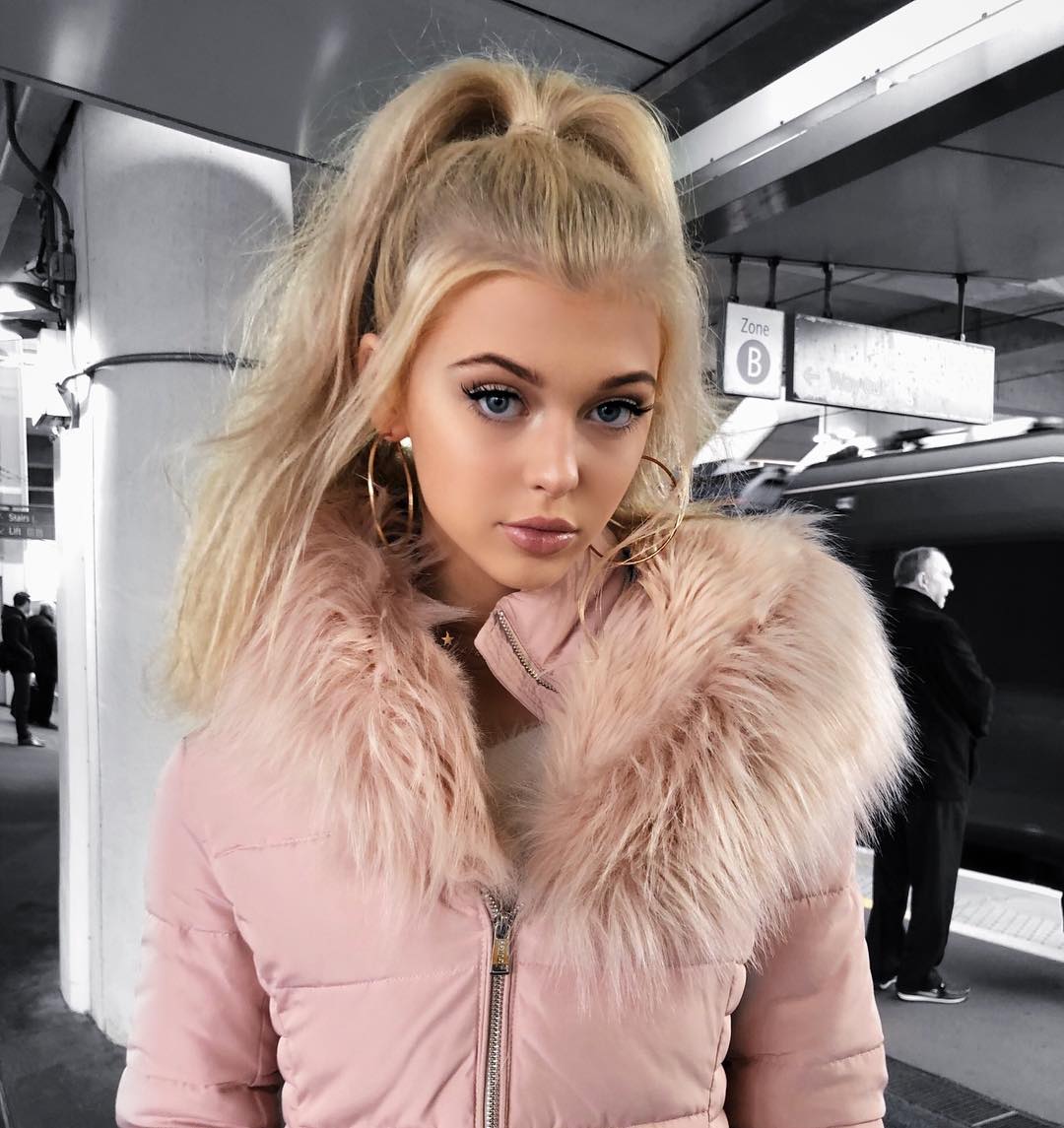 a young woman with blonde hair and blue eyes is posing for a photo in a subway station. she is wearing a pink puffer jacket with a fur collar and large hoop earrings. her hair is styled in a high ponytail. in the background, there is a subway train, a "zone b" sign, and a couple of people waiting. the overall color scheme is muted, with pink as the dominant color.