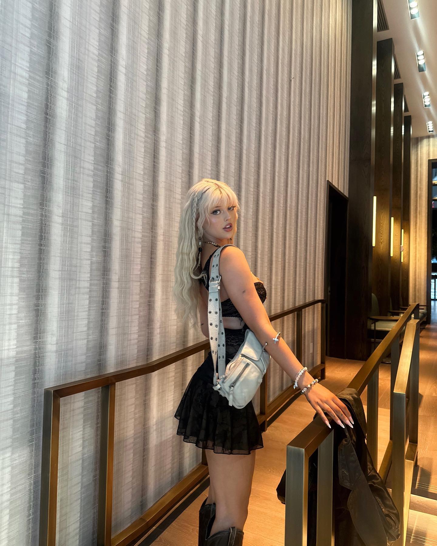 a woman with blonde hair and a black outfit is posing indoors near a railing. she is wearing a skirt and boots, and carrying a white bag. she also has jewelry on her wrist and neck. the background is a hallway with architectural details.