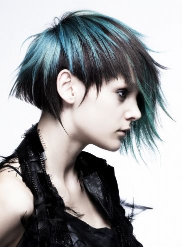 a woman with a blue and black, edgy, asymmetrical haircut poses against a white background in a studio setting.  she has dark makeup and is wearing a black, feathered top.  the lighting is bright and highlights the details of her hair and the texture of her clothing.