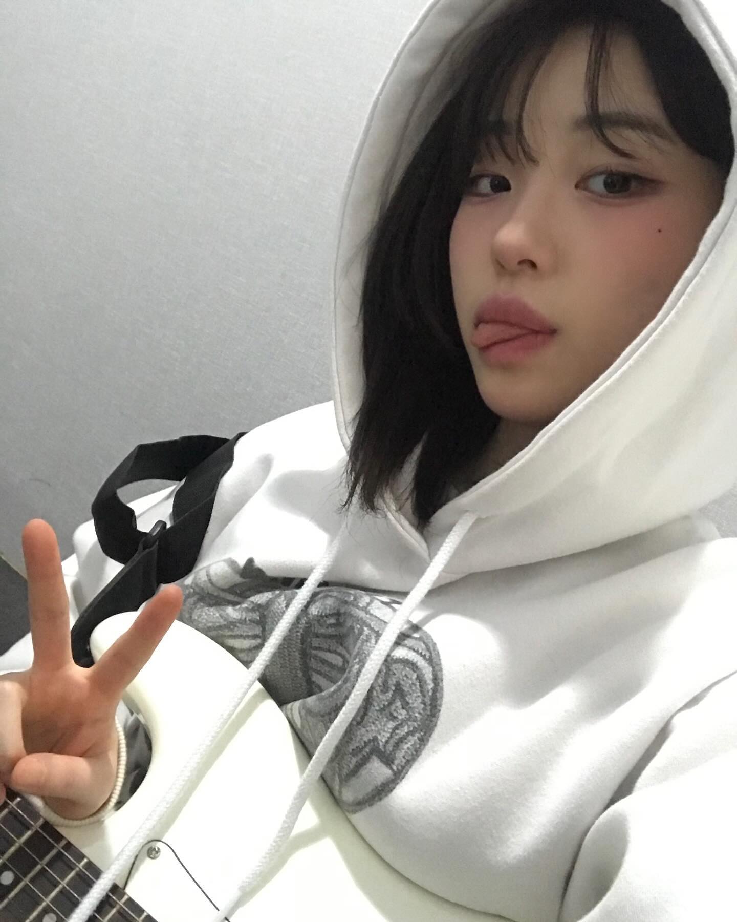 a young woman poses for a selfie while holding a white electric guitar. she's wearing a white hoodie with the hood up and is making a peace sign with her left hand. she has short, dark hair and is sticking her tongue out playfully. the setting appears to be an indoor studio with bright, artificial lighting.