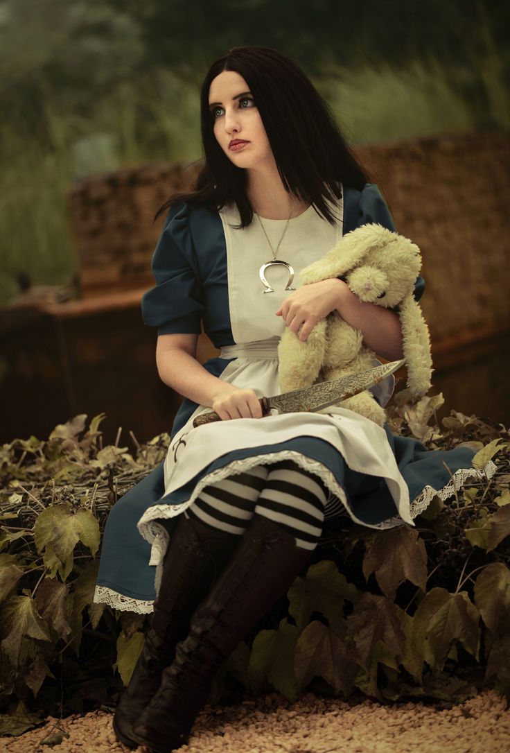 a young woman in a meticulously crafted alice in wonderland cosplay sits pensively outdoors. she is dressed in a dark blue dress with a white apron, striped stockings, and tall boots. she holds a stuffed bunny and a knife, adding a touch of darkness to the classic character.  the setting appears somewhat overgrown and mysterious, enhancing the overall gothic and fantasy feel. the cosplay is highly detailed, and the model's expression is thoughtful and slightly melancholic.  the omega symbol on her necklace suggests a possible deeper interpretation of the character, perhaps drawing inspiration from video game adaptations.