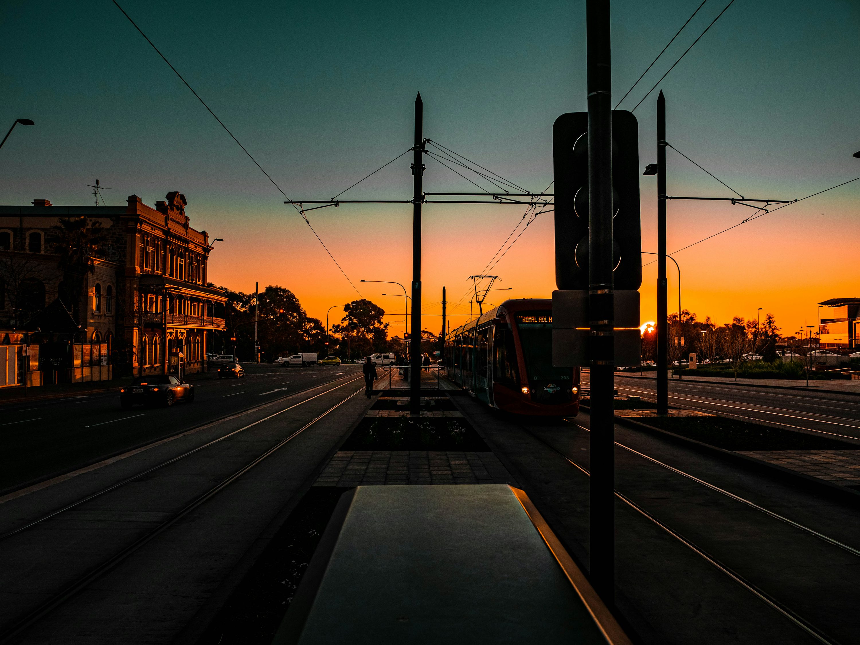 a cityscape view at sunset featuring a tram on its tracks, with architecture and infrastructure elements including traffic lights and buildings against an orange and blue sky.
