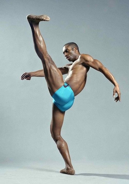 a young male dancer in blue briefs is in a powerful lunge pose in a studio with a gray background.  his leg is stretched out at a 90 degree angle in front of him with his toes pointed, he is looking off to the side.  his muscles are flexed and he looks very strong and powerful.