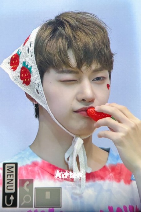 a closeup selfie of a young asian man wearing a white crocheted strawberry hat. he is winking at the camera while holding a red strawberry to his lips and wearing a colorful tshirt. the background is a light blue.