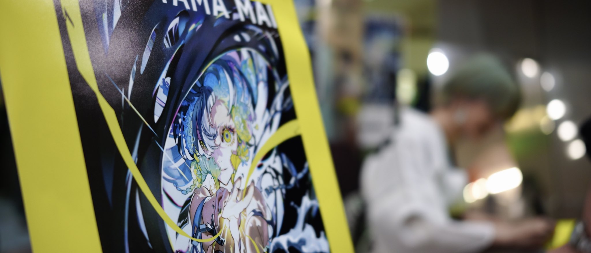 a closeup shot of a poster featuring an animestyle illustration. the poster has a yellow border and showcases a character with blue hair and intricate details. the background is blurred, but a person wearing a green cap is visible, seemingly working on something under bright, artificial lighting. the overall setting suggests an indoor environment, possibly a convention or an artist's workspace.
