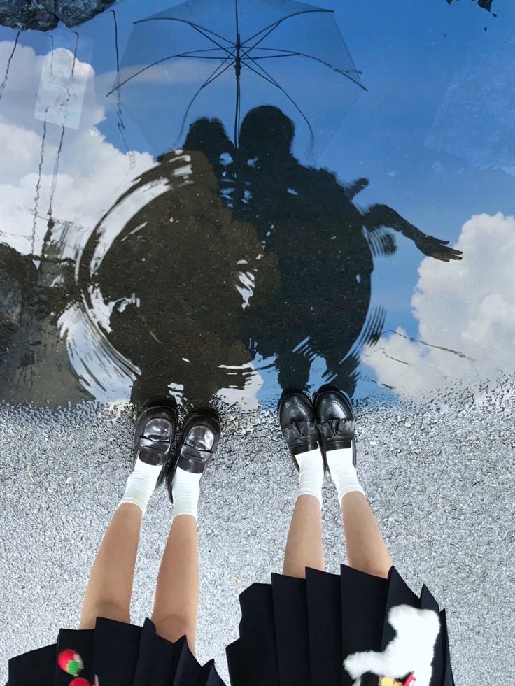 a topdown shot of two girls in school uniform standing in a puddle. their reflection is visible in the puddle, showing them with an umbrella. the sky is partly cloudy.  the girls are wearing black loafers and white socks.