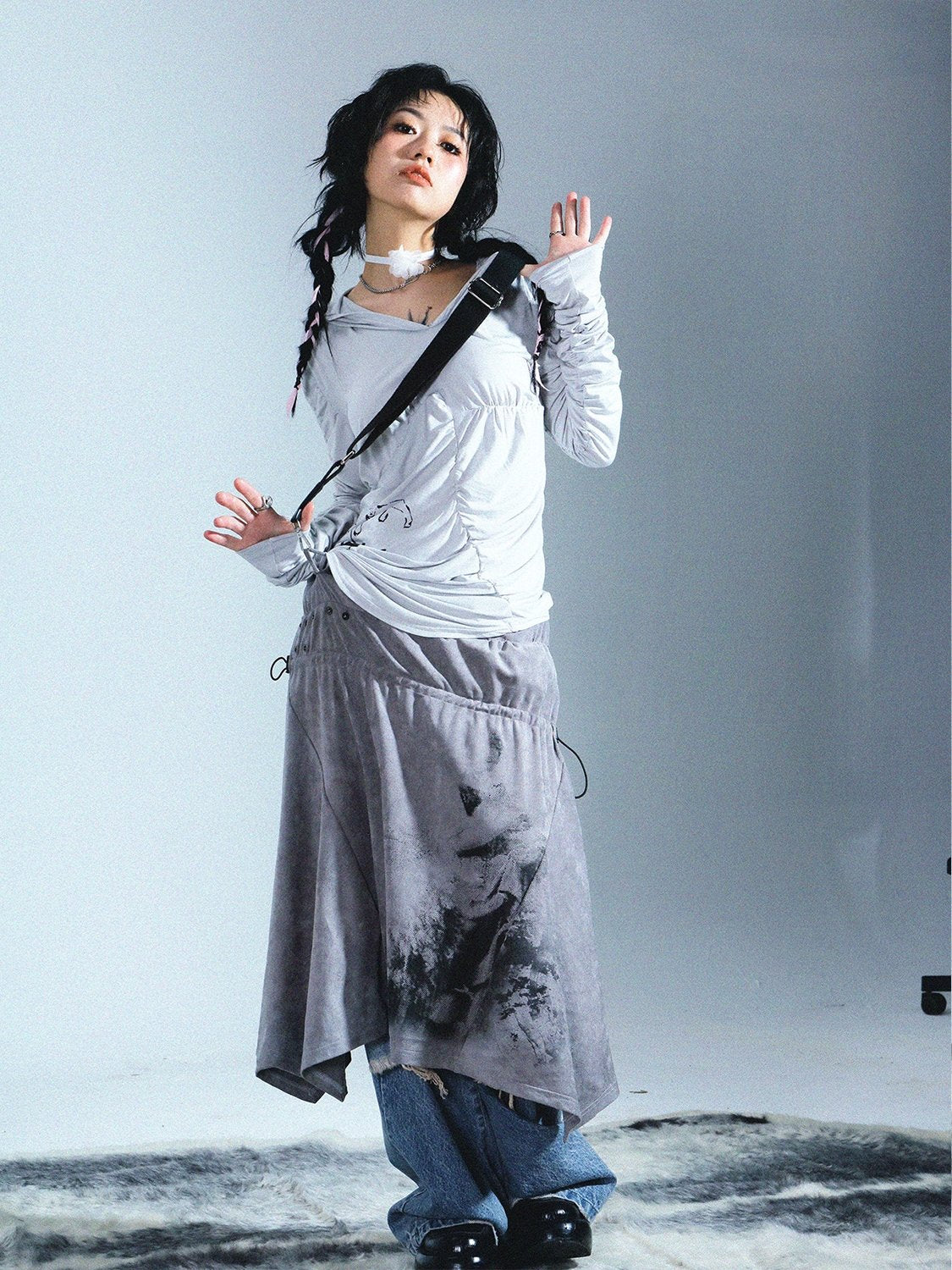 a fulllength studio shot depicts a young woman modeling a stylish and slightly grungeinspired outfit. she's wearing a light grey, loosefitting, longsleeved top with an asymmetrical hem. the top is paired with a grey asymmetrical skirt featuring a dark, artistic print.  the skirt is long and drapes loosely. underneath the skirt, distressed blue jeans are visible at the ankles. she accessorizes with a simple black bag strap slung across her shoulder and a delicate choker. her dark hair is styled in loose pigtails.  the background is a minimalist light grey, drawing focus to the model's attire and pose. the overall style suggests a blend of streetwear and artistic flair.