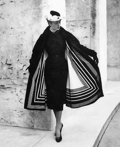 a black and white fulllength portrait of a woman in a stylish outfit. she is wearing a black dress, a black coat with a striking black and white striped lining, a hat, gloves, a pearl necklace, and heels. she stands outdoors against a textured wall and a stone ledge. the photograph exudes a vintage and elegant aesthetic.