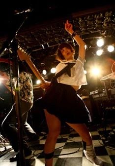 a young woman, possibly a japanese musician, is captured midperformance on stage.  she's wearing a sailorstyle school uniform skirt and is energetically playing an electric guitar. her pose is dynamic, suggesting an intense and exciting musical moment. the background is dark and features concert lighting, indicating a live show in a club or small venue. a partially visible second person is also playing a guitar in the background. the floor is a black and white checkered pattern. the overall atmosphere is one of vibrant energy and youthful rebellion.