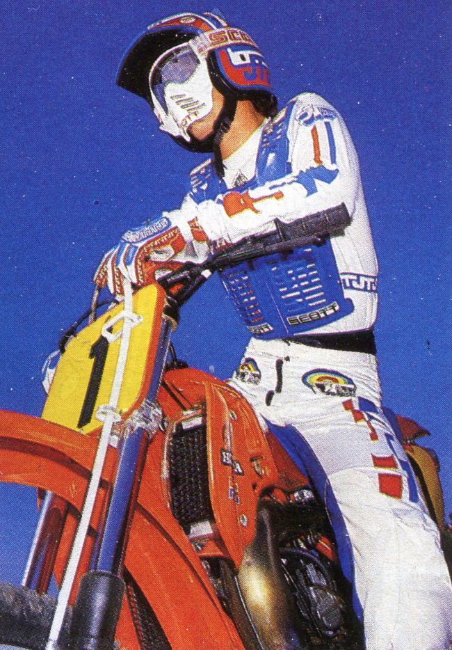 this image captures a motocross rider in action. the rider is wearing a white and blue racing suit, a fullface helmet with goggles, and gloves. the rider is sitting on a red and yellow dirt bike with a "1" race number plate. the rider's arms are extended, showing off their powerful stance and control as they navigate a dirt track. the blue sky and the vibrant colors of the rider's gear create a dynamic and energetic scene, reflecting the excitement and thrill of motocross racing.