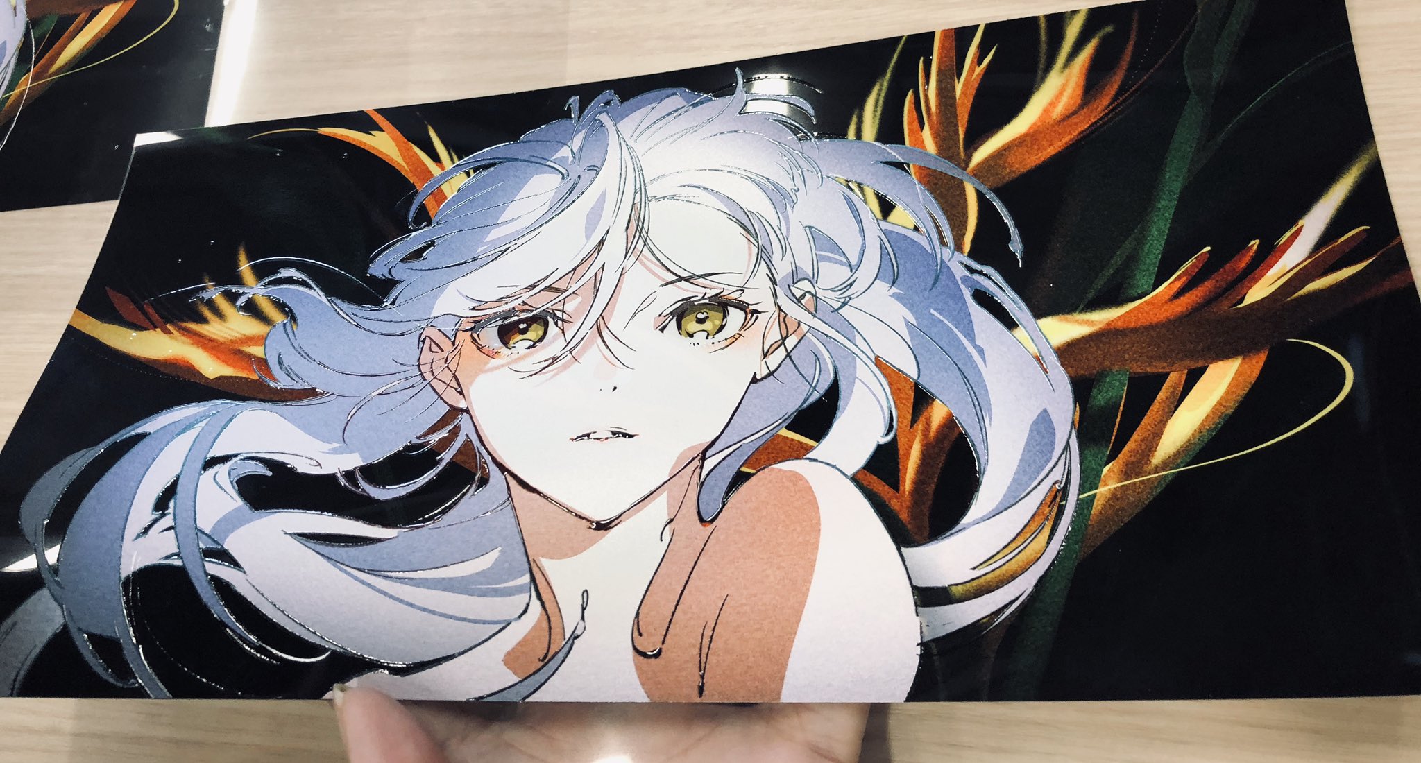 the image shows a print of a digital illustration featuring an animestyle girl with long, flowing white hair and striking yellow eyes. she's set against a dark backdrop with vibrant orange and green bird of paradise flowers, creating a stark yet harmonious contrast. the perspective is a closeup, emphasizing her delicate features and the overall ethereal feel of the artwork. the print itself has a glossy finish.
