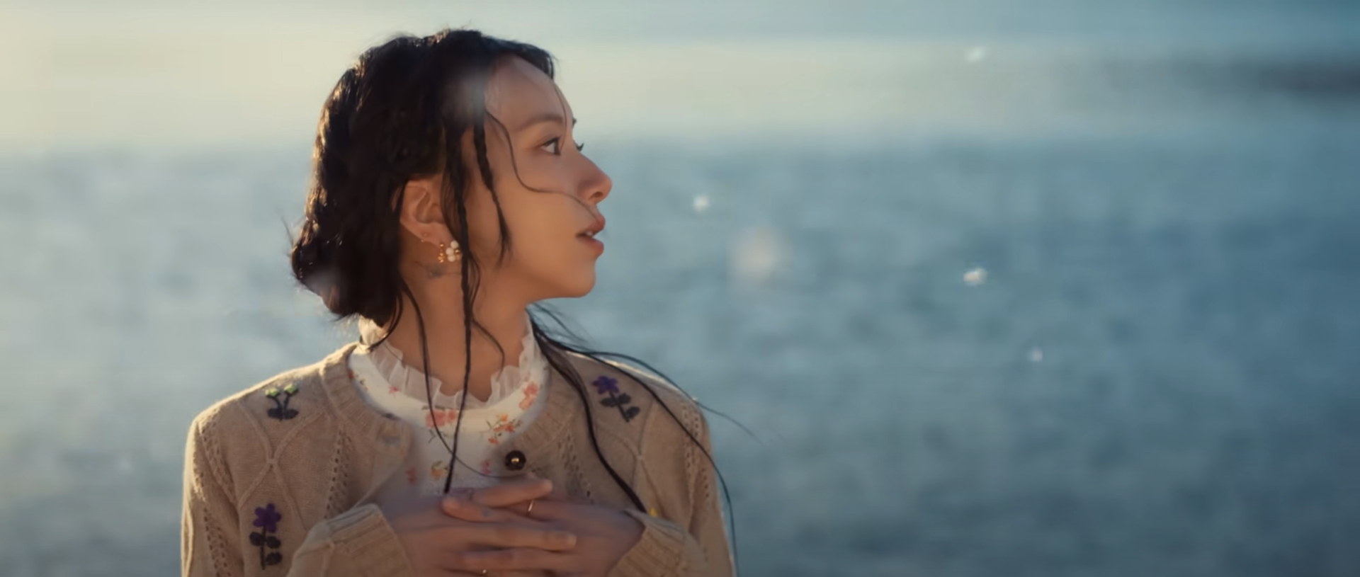 a young woman with long dark hair stands on a snowy beach, looking towards the right side of the image. the sun is setting, casting a warm, soft light on the scene. the woman is wearing a beige cardigan with floral embroidery. her expression is thoughtful and introspective, suggesting a sense of peace and contemplation. the background is a blurry blend of snow and water, creating a serene and ethereal atmosphere.