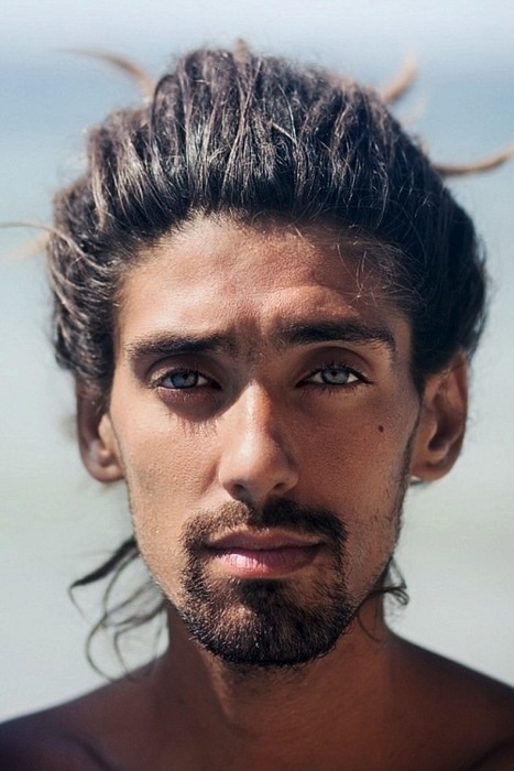 a closeup portrait of a handsome man with dark hair pulled back into a messy bun or ponytail. he has light skin, piercing blue eyes, a neatly trimmed beard and mustache, and is gazing directly at the camera. the lighting is soft and natural.