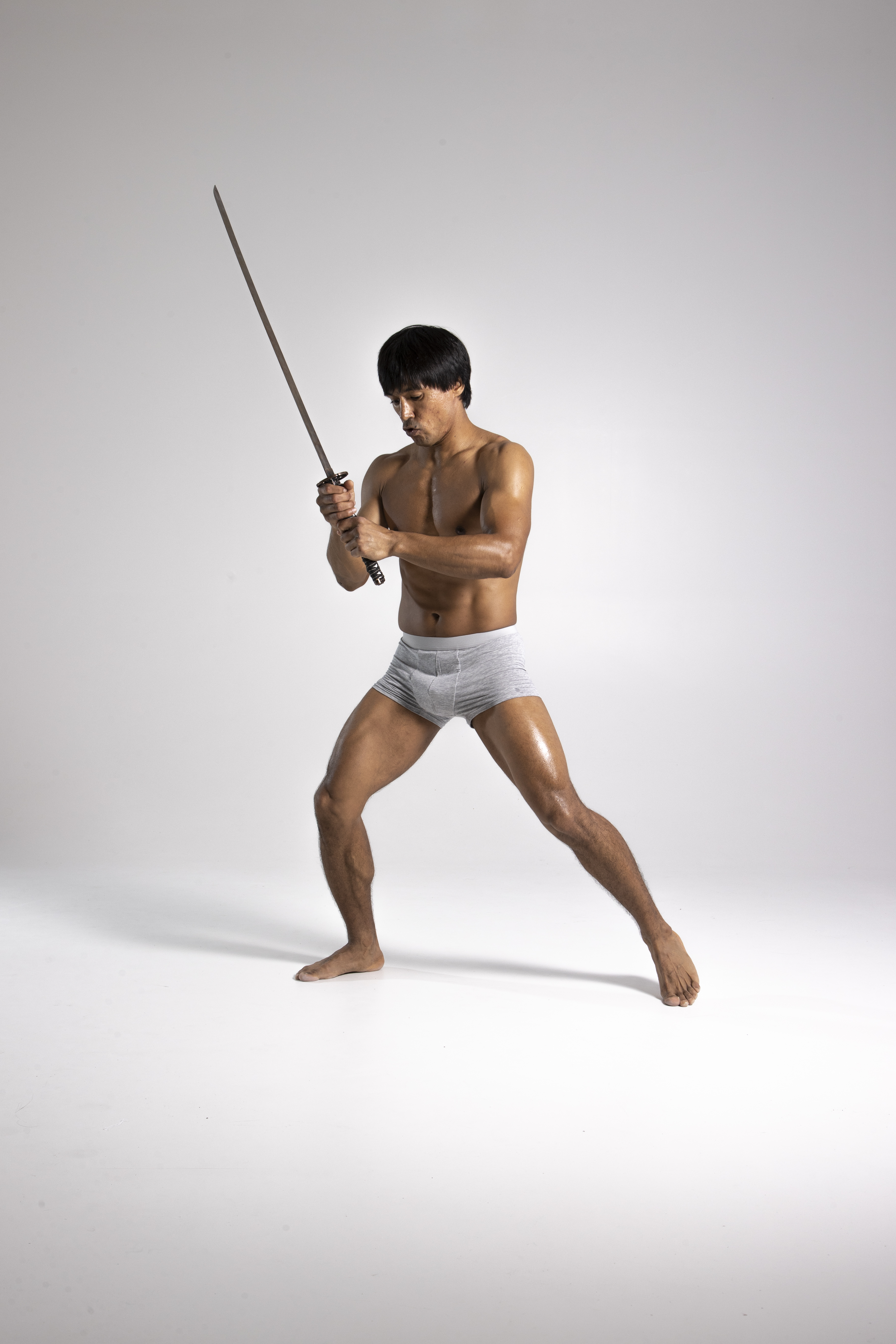 a shirtless asian man in underwear holds a katana above his head in a martial arts pose. he is muscular and in a studio setting with a white background.