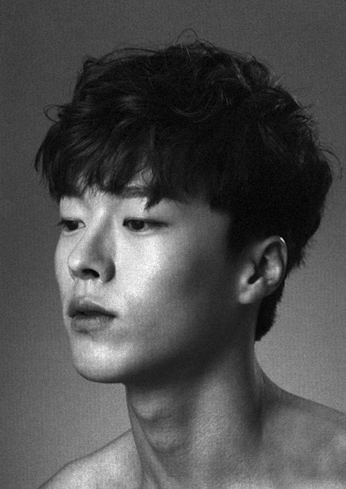 the image is a black and white, closeup portrait of a young asian man.  he has short, slightly tousled dark hair and is looking down and to the side, creating a pensive or contemplative mood. his expression is serious and his chest and shoulders are bare. the background is a plain, muted gray, focusing all attention on the subject. the lighting is dramatic, adding depth and shadow to his features. the overall style suggests a fine art photograph or a fashionstyle portrait.