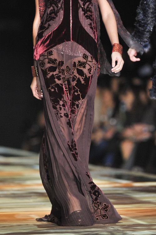a model walks down a runway wearing a long, black velvet gown with floral embroidery and sheer panels. the dress has a fitted bodice and a flowing skirt. the model is wearing delicate bracelets on both wrists and has her hair styled in a sleek updo. she is posing for the camera as she walks down the runway.