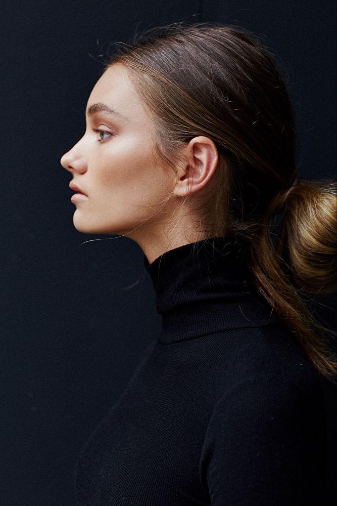 a side profile portrait of a young woman with brunette hair styled in a low bun. she is wearing a black turtleneck sweater and standing against a dark background. the lighting is soft and natural.