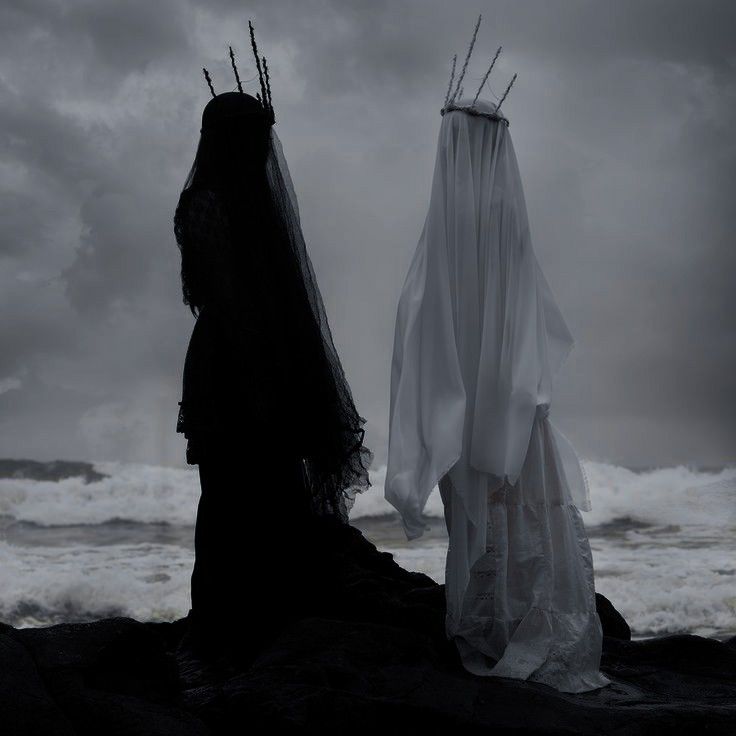 two women stand on a rocky beach facing the stormy ocean. one woman is dressed in black with a crown and veil covering her face, while the other woman is dressed in white with a crown and veil, making her look like a ghost. the dark and light imagery creates a stark contrast, while the stormy ocean and dramatic lighting contribute to the mysterious and gothic atmosphere.
