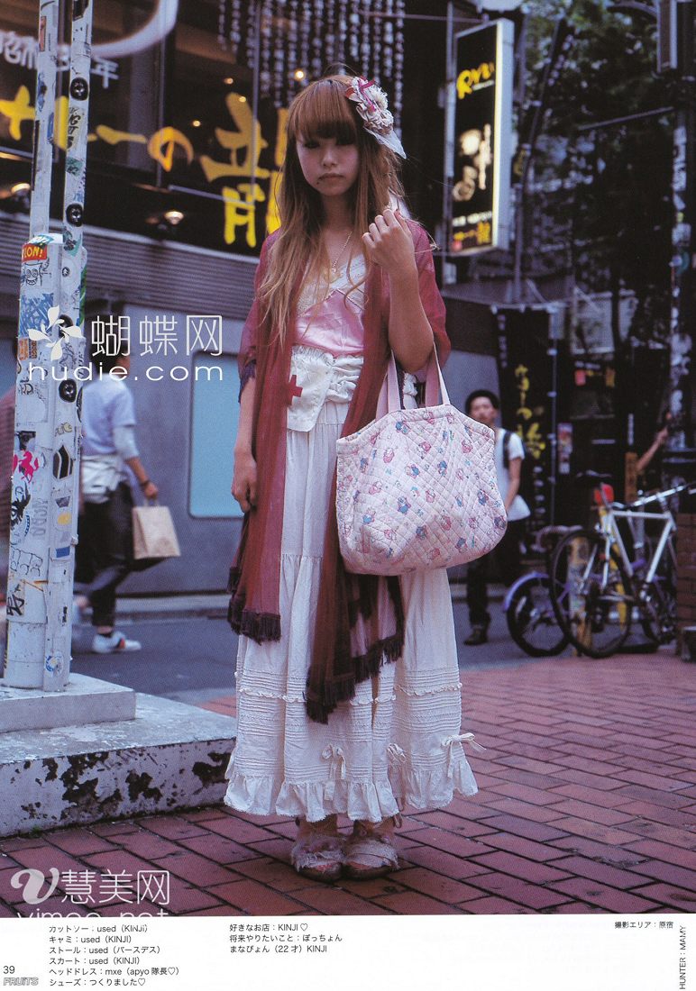 a young woman is posing on a city street, her outfit a mix of vintage and modern styles. she wears a long white tiered skirt, a pink corset top, and a deep red shawl. her hair is long and straight, with bangs framing her face. she carries a large floral tote bag. the background is a busy japanese street scene, with storefronts, bicycles, and other pedestrians. the image appears to be from a fashion magazine, as indicated by the japanese text and website address superimposed on the photo.