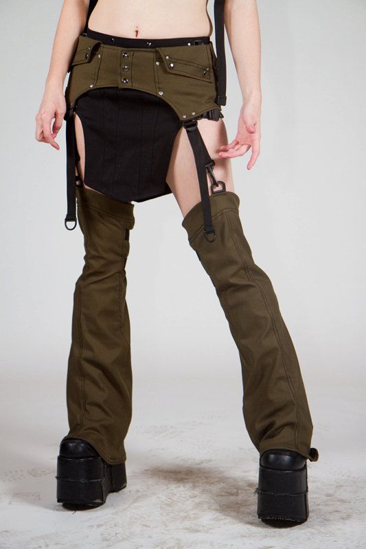 a young woman posing in a studio wearing a black and green fashion ensemble. the woman is wearing black boots, green pants, and a black skirt with a green belt. she is wearing a lot of straps and buckles.