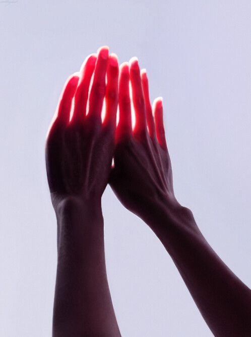 a closeup image of a person's hands, illuminated from below so the light shines through their skin and makes their fingers look red. the hands are clasped together and the wrists are slightly bent.  the background is a simple, light gray.