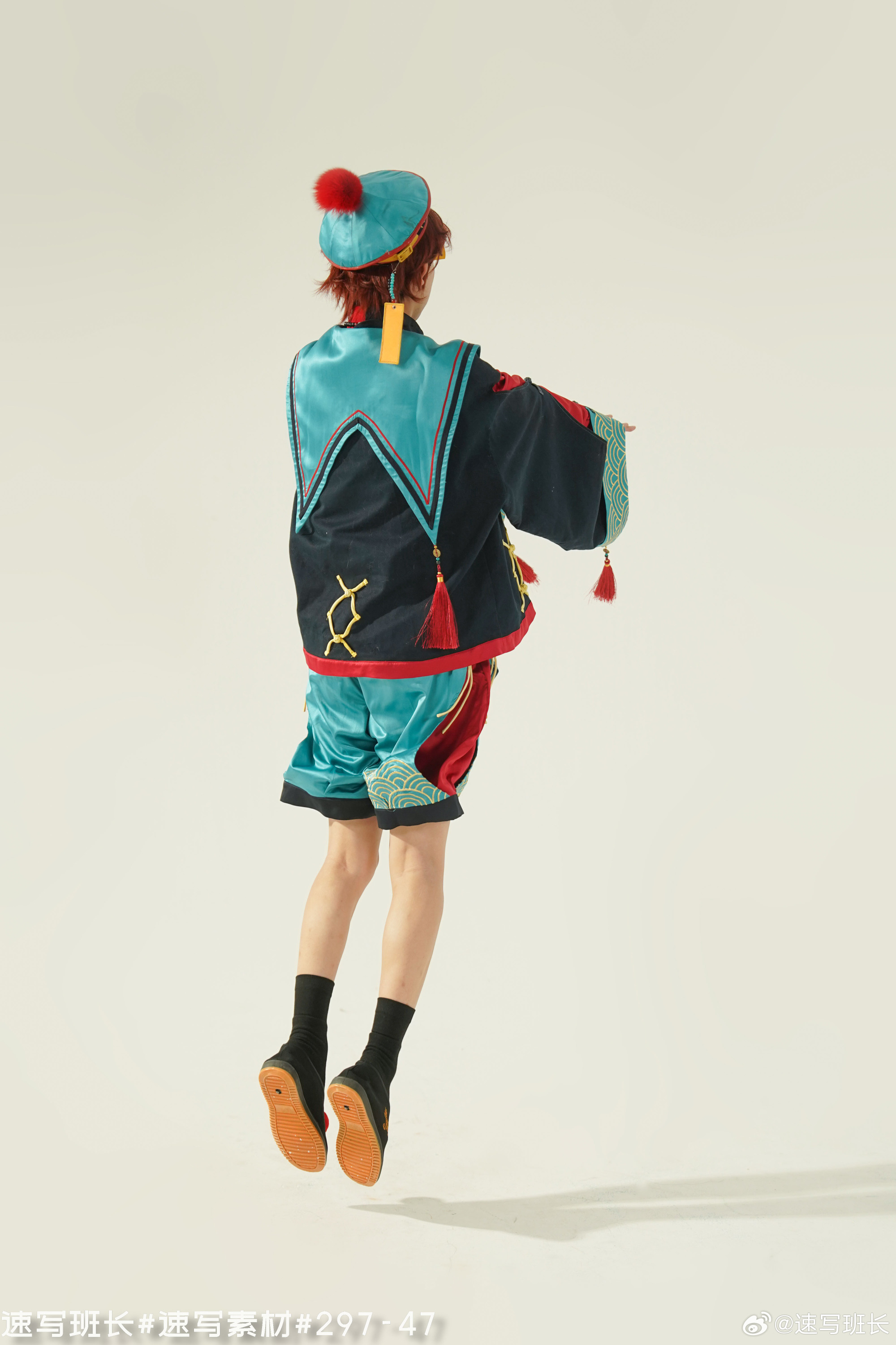 a young male model is captured midair, executing a jump while facing away from the camera. he is adorned in a vibrant, modernized rendition of hanfu, a style of traditional chinese clothing. the outfit features a predominantly blue and black color scheme, accented with red and gold details. the top is a short, kimonolike jacket with wide sleeves, while the bottom consists of loosefitting shorts.  his ensemble is further enhanced by a matching hat with a red pompom, completing the eyecatching attire. the backdrop is a minimalist white studio setup, drawing focus to the model and his dynamic pose. the studio lighting illuminates the scene, casting subtle shadows that accentuate the textures and folds of the clothing.