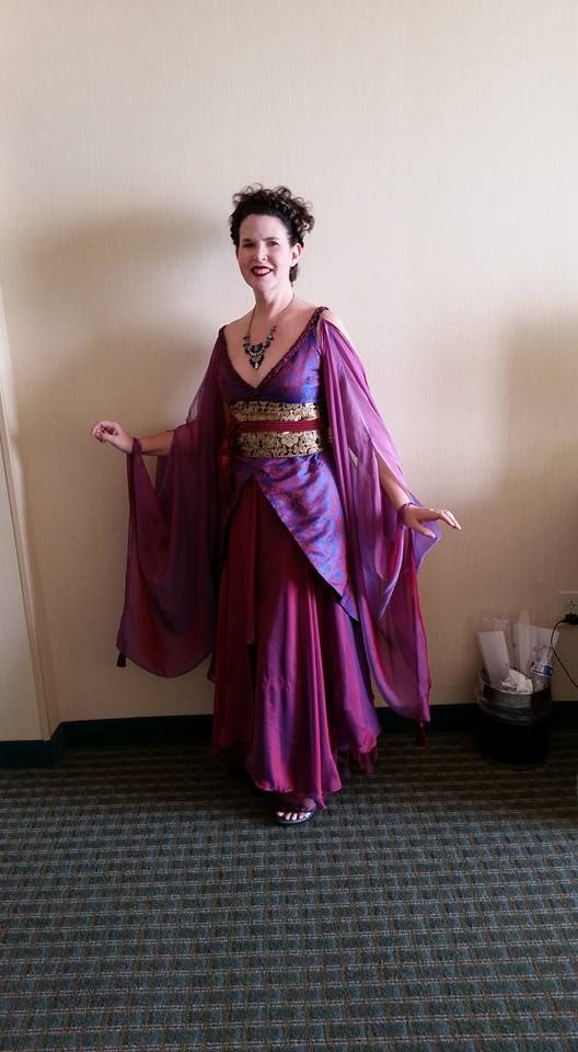 a woman is standing in a long purple gown with flowing sleeves. the dress has a gold belt and she is wearing a necklace. she is smiling and appears to be posing for a photo. the setting appears to be indoors on a carpeted floor. the style of the dress suggests a fantasy or cosplay costume.