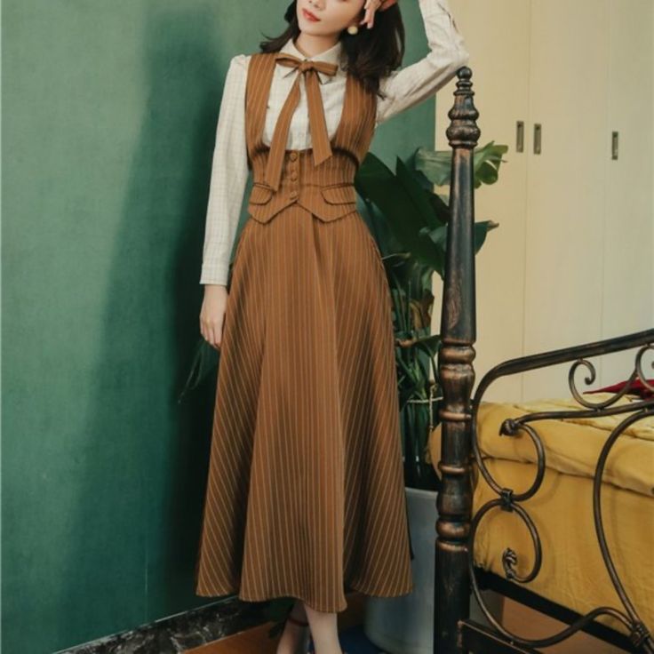woman is standing in an indoor setting wearing a vintagestyle brown dress with vertical stripes. the dress is midi length and has a highwaisted design with a sleeveless top. she is also wearing a white longsleeved blouse with a bow tie. the background features a green wall and a vintagestyle bed. the lighting is artificial.