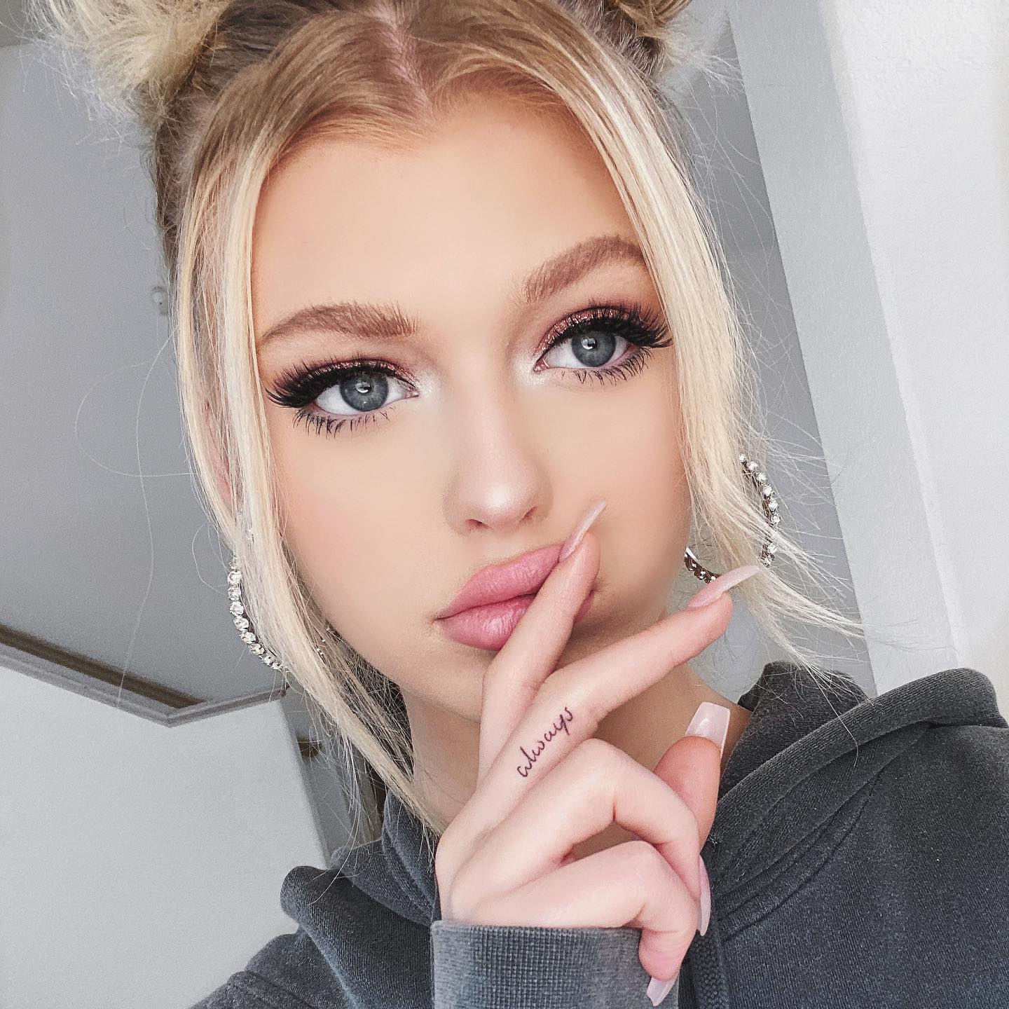 a closeup portrait of a young woman with blonde hair styled in two buns on top of her head. she has long nails, pronounced eye makeup, and is wearing hoop earrings. a tattoo is visible on the side of her index finger. she is wearing a gray hoodie.